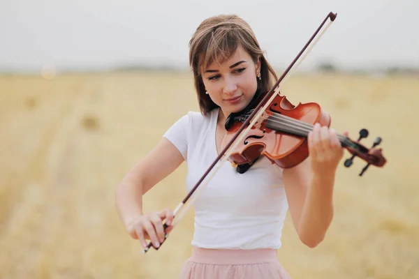 Beautiful romantic girl with loose hair playing the violin in the field after the harvest. Square sheaves of hay in the field. Violin training