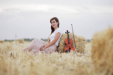 Saçları dalgalanan romantik genç bir kadın, hasattan sonra elinde bir kemanla bir tarlada. Tarlada kare demet saman. Keman eğitimi