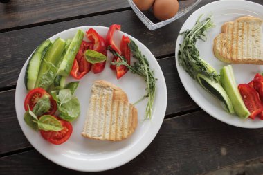 Sağlıklı organik sebze kahvaltısı, domates, tost, salata, fesleğen, yumurta. Siyah ahşap bir masada beyaz tabakta vejetaryen sağlıklı yiyecekler. Yüksek kalite fotoğraf