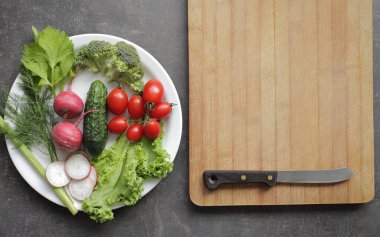Gri bir masada beyaz tabakta taze sebzeler. Domates, salatalık, marul, brokoli, turp, kesim tahtası.