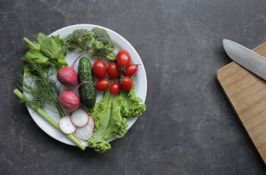 Gri bir masada beyaz tabakta taze sebzeler. Domates, salatalık, marul, brokoli, turp, kesim tahtası.