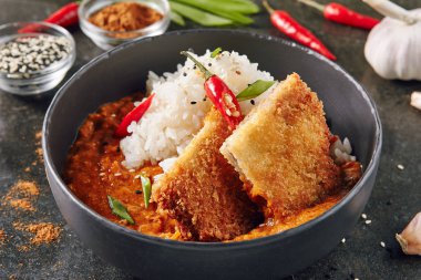 Köri ve pilav koyu arka plan üzerinde sıcak baharatlı çıtır çıtır kızarmış tavuk fileto. Katsu veya Tonkatsu kırmızı Peppres, Türkiye'de et pirzola, ekmek kırıntıları, domates, biber sosu, susam, yeşillik ve otlar ile