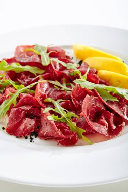 Kurutulmuş İtalyan sığır Bresaola, limon, taze fesleğen, roka ve balsamik krem beyaz arka plan üzerinde izole. Air-dried ve tuzlu Dana, at, geyik eti veya domuz eti ile Yeşiller düz Restoran plaka üzerinde