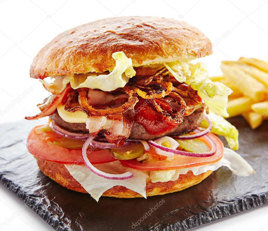 Fresh Beef Burger with Bacon, Fried Onions, Tomato Sauce, Pickled Cucumbers, Green Lettuce, Cheese and French Fries Garnish. Hamburger or Beefburger on Natural Black Stone Plate Isolated on White