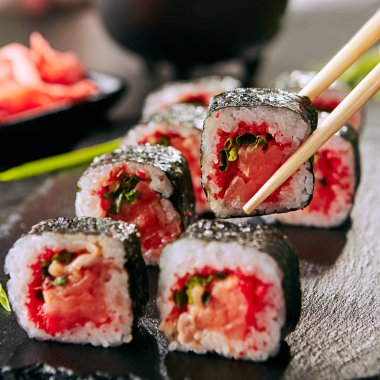 Chopstick yeşil soğan, uçan balık kırmızı havyar, baharatlı sos ve bulanık arka plan üzerinde nori ile füme somon suşi rulo tutan. Siyah kayrak plaka arka plan closeup taze gurme Japon gıda