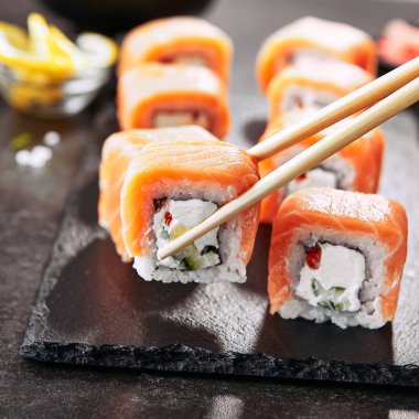 Chopstick Siyah Kayrak Plaka Close Up üzerinde Somon ve Krem Peynir ve Cuccumber ile Sushi Rolls tutan. Uramaki, Nori Maki veya Futomaki Suşi Alabalık Fileto, Soya Sosu ve Wasabi ile