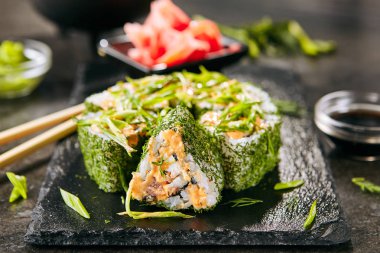 Pirinç, krem peynir, kızarmış somon, domates, pekin lahanası, yeşil soğan, dereotu ve baharatlı sos closeup ile Yeşil uramaki suşi rulo. Siyah kayrak arka plan üzerinde renkli maki susi rulo makro çekim