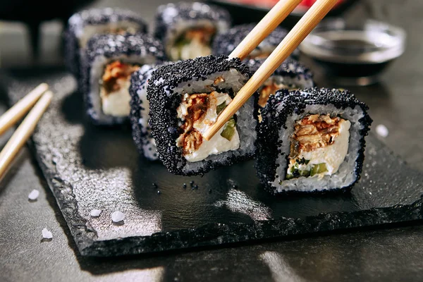 クリームチーズ うなぎ きゅうり 飛ぶ魚の黒キャビアと海苔で巻くうなぎ巻き箸 黒いスレートプレートの背景にトビコと新鮮な日本のウラマキロール — ストック写真