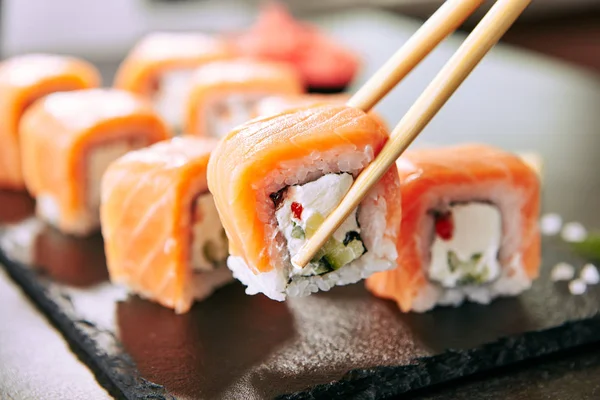 筷子持有寿司卷设置与鲑鱼和奶油奶酪和凝固在黑色石板板特写 乌拉马基 诺里马基或富托马基寿司与鳟鱼片 酱油和瓦萨比 — 图库照片
