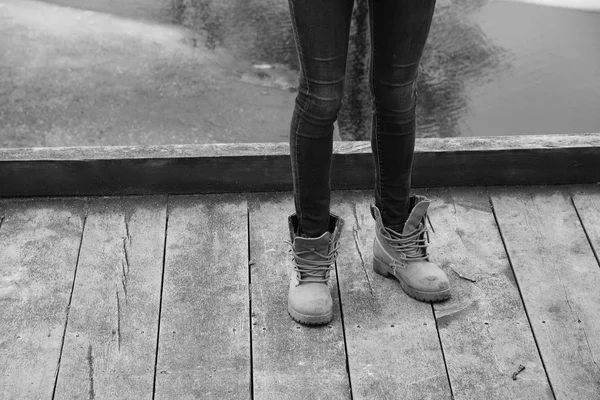 Young person in tight jeans and untied shoes standing on wooden pier.