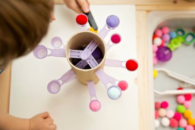 Evindeymiş gibi davran. Koordinasyon, denge ve motor becerileri geliştirmek için DIY oyuncağı. Çocuk, tatlı kaşıklarına ponpon yerleştiriyor..