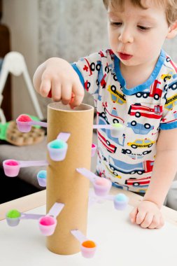 Evindeymiş gibi davran. Koordinasyon, denge ve motor becerileri geliştirmek için DIY oyuncağı. Çocuk, tatlı kaşıklarına ponpon yerleştiriyor..