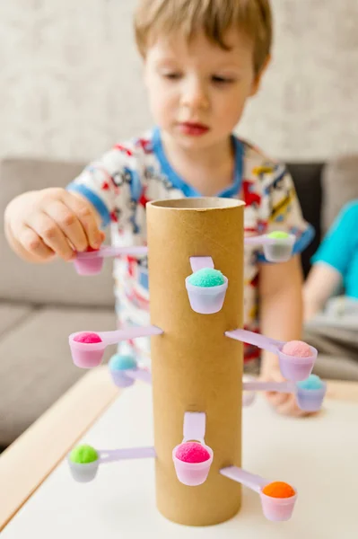 Evindeymiş gibi davran. Koordinasyon, denge ve motor becerileri geliştirmek için DIY oyuncağı. Çocuk, tatlı kaşıklarına ponpon yerleştiriyor..