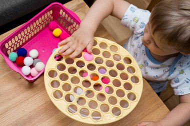 Odaklanmış, konsantre bebek yüzü. 2 yaşında bir çocuk deliğe ponponları sokarak Rus Dumpling Maker 'la oynuyor. Plastik sepette renkli iplik topları