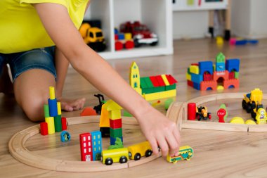 Kulaklıklı çocuk yerde tahta oyuncaklarla oynuyor. Tren rayları, ahşap blok binalar.