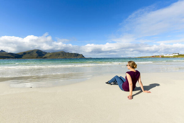 Beautiful young blond woman in dress is sitting on the sand of caribbean beach near azure sea at sunset