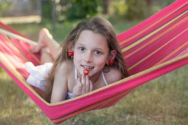 Kırmızı bir hamak beyaz elbiseli bir kız yatıyor. Kız çilek, hastası kiraz. Bir kız bir kiraz ile onun elinde gülümsüyor.