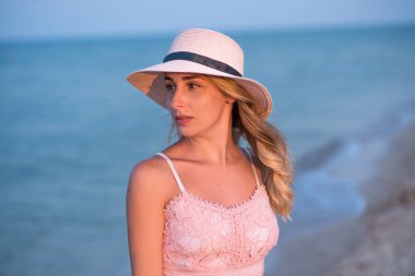 Deniz kenarında tatilde olan genç bir kız. Denizde gün batımı. Elbiseli, şapkalı ve sahilde yürüyen mutlu genç bir kadın. Aile bağlamında. Seyahat et ve rahatla