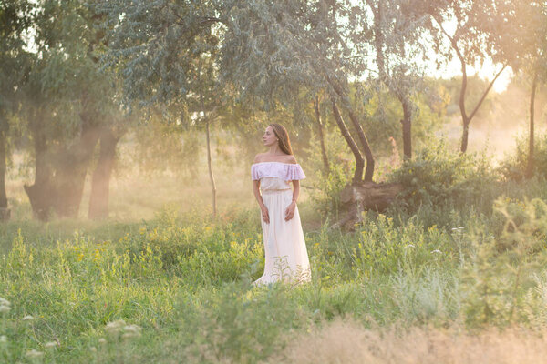 Young woman walking in the park. Beautiful girl in a long dress at sunset. Summer and relaxation. Travels