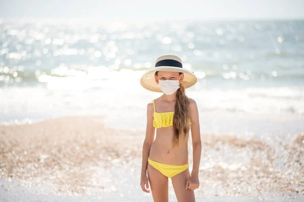 Beautiful girl alone on the beach in a medical mask. A crisis. How to wear a mask when traveling. Family content. Child isolated on the beach. Keep your distance. Beautiful long-haired girl in a swimsuit on the ocean