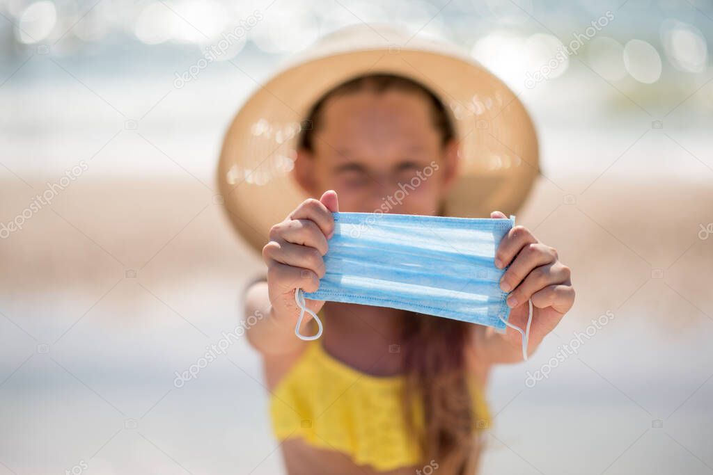 A child on the beach puts on a medical mask. Coronavirus and crisis. It is safe to spend time on vacation. Beautiful girl with long hair in a yellow swimsuit by the ocean. Traveling during a pandemic.