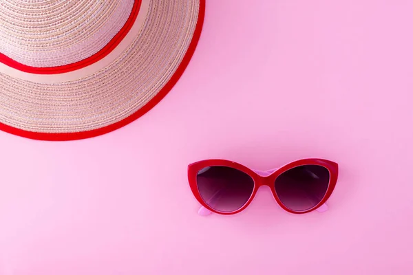 A red sunglasses and bright beach hat on a light pink background.