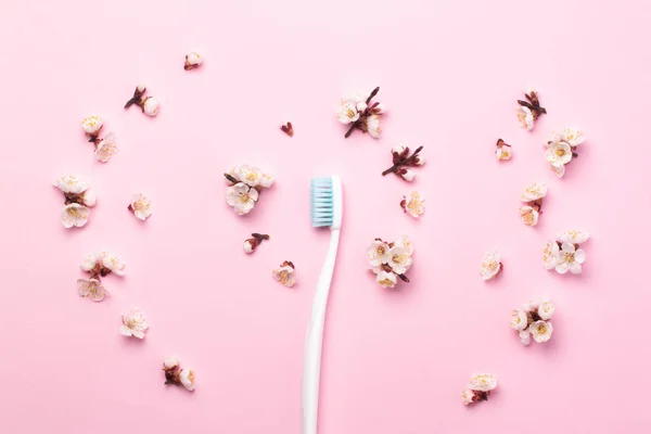 แปรงสีฟันสีขาวที่มีขนสีฟ้าบนพื้นหลังกระดาษสีชมพูตกแต่งด้วยดอกไม้ฤดูใบไม้ผลิ . — ภาพถ่ายสต็อก