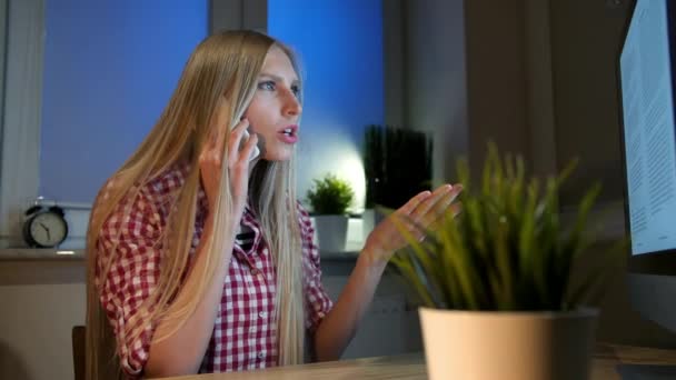 Donna stupita a computer che parla su smartphone. Attraente donna bionda seduta alla scrivania di legno e guardando a bocca aperta il monitor del computer che parla sul telefono cellulare in camera oscura di notte — Video Stock