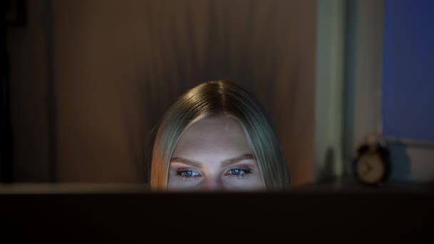 여자가 밤에 컴퓨터 모니터를 훔쳐 보고 있어요. 밤에 컴퓨터 앞에 앉아 있는 아름다운 여성의 모습 과 희미 한 배경 위에 검은 창 이 있는 모니터 위에 있는 카메라를 보고 있는 모습 — 비디오