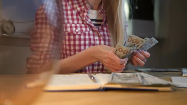 Mujer contando dinero en efectivo en el escritorio. Atractiva hembra rubia con camisa a cuadros sentada por la noche en la habitación con mesa de madera con lámpara y computadora y contando fajos de billetes en las manos . — Vídeos de Stock