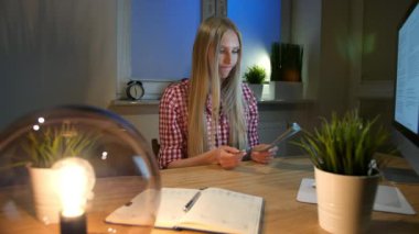 Kadın holding bitcoins ve düşünme. Güzel sarışın kadın ile bilgisayar masası, karanlık oda ve gece lambası oturan ve elinde tutan bitcoins yansıtan.