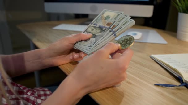Egy nő bitcoinnal és egy köteg pénzzel. Növényi kilátás női kéz kezében fém bitcoin és köteg dollár bankjegyek ül fa íróasztal jegyzettömbbel és számítógép monitor éjjel. — Stock videók