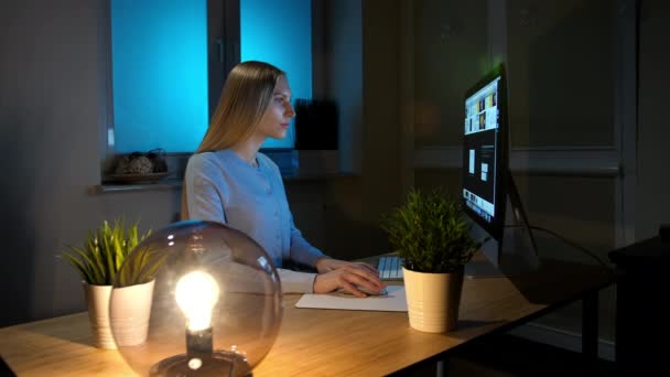 Mujer mirando atentamente a la computadora por la noche. Relajada hermosa rubia en ropa de hogar caliente sentada en la mesa de madera iluminada por una pequeña lámpara de esfera y concentrada en la navegación en el ordenador . — Vídeo de stock