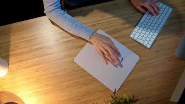 Una donna che lavora al computer di notte. Crop vista delle mani della donna in comodi vestiti di casa seduto alla scrivania di legno acceso dalla lampada e utilizzando il mouse del computer e la tastiera in camera oscura di notte . — Video Stock