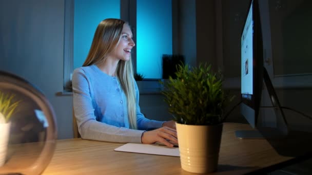Χαμογελαστή γυναίκα που δουλεύει στον υπολογιστή τη νύχτα. Χαμογελαστή γυναίκα με καρό πουκάμισο που κάθεται στο φως από μικρό λαμπτήρα ξύλινο γραφείο και κοιτάζοντας με ενθουσιασμό στην οθόνη του υπολογιστή γράφοντας πληροφορίες. — Αρχείο Βίντεο