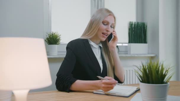 Zakenvrouw aan het praten op smartphone in het kantoor. Elegante jonge blonde vrouw in kantoorpak die op het werk zit en onderhandelt via mobiele telefoon in de hand en de nodige informatie opschrijft in het notitieboekje — Stockvideo