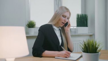 Ofiste akıllı telefondan konuşan bir iş kadını. Ofisteki zarif sarışın kadın iş yerinde oturuyor ve elinde cep telefonuyla gerekli bilgileri not defterine yazıyor.