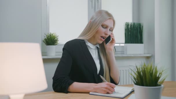 Zakenvrouw aan het praten op smartphone in het kantoor. Elegante jonge blonde vrouw in kantoorpak die op het werk zit en onderhandelt via mobiele telefoon in de hand en de nodige informatie opschrijft in het notitieboekje — Stockvideo