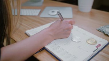 Günlük ajandasında bitcoin yazan bir kadın. Günlük planlayıcıyla kalem yazma verisi olan, sayfasındaki bitmadeni paralarla açık ahşap masaya yerleştirilmiş bir kadın görüntüsü.
