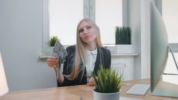 Una mujer relajada con dinero en la oficina. Elegante joven rubia en traje de negocios sentada en el escritorio con computadora y planta y sosteniendo en la mano ventilador de efectivo con confianza mirando a la cámara . — Vídeos de Stock