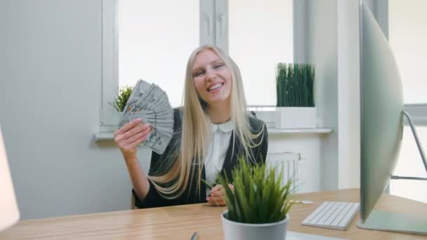 Усміхнена ділова жінка тримає кусок готівки. Красива щаслива блондинка сидить на робочому місці з комп'ютером і тримає шанувальника валютних банкнот і дивиться на камеру посміхається . — стокове відео