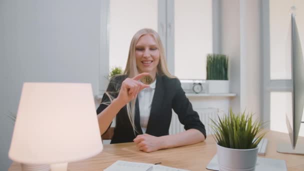Happy female with bitcoin showing thumbs up. Smiling cheerful blond woman in office suit sitting at workplace with computer and showing bitcoin in hand doing thumbs up gesture and looking at camera — Stock Video