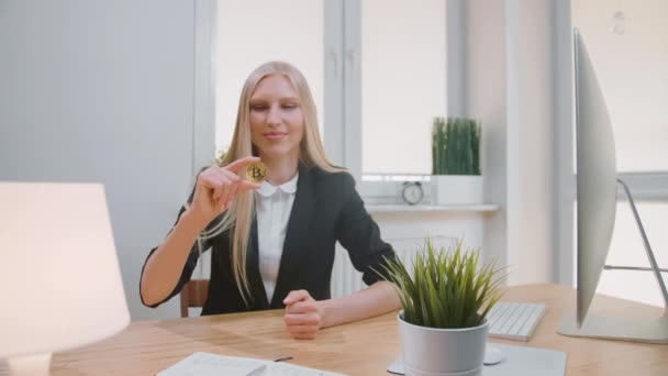 Счастливая женщина с биткойном, показывающая большие пальцы вверх. Улыбающаяся светловолосая блондинка в офисном костюме, сидящая на рабочем месте с компьютером и показывающая биткойн в руке, делая большой жест и глядя в камеру — стоковое видео