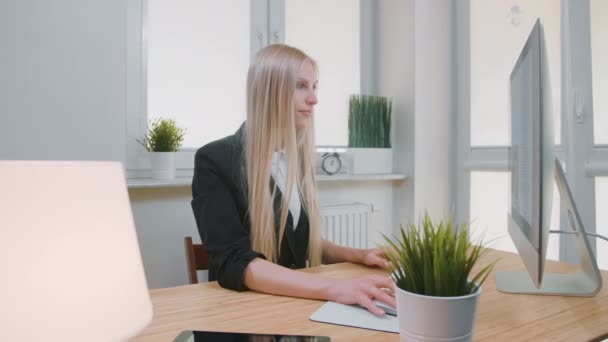 En leende kvinna som jobbar på en dator på kontoret. Vacker ung blond kvinna sitter på arbetsplatsen med dator och håller hand på mus tittar på kameran och ler. — Stockvideo