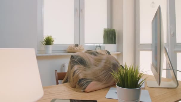 Уставшая женщина просыпается на работе. Уставшая блондинка-офисный работник в элегантном костюме расслабляется лежа на руках на столе, затем встает зевая и начинает работать за компьютером в светлой комнате с растениями . — стоковое видео
