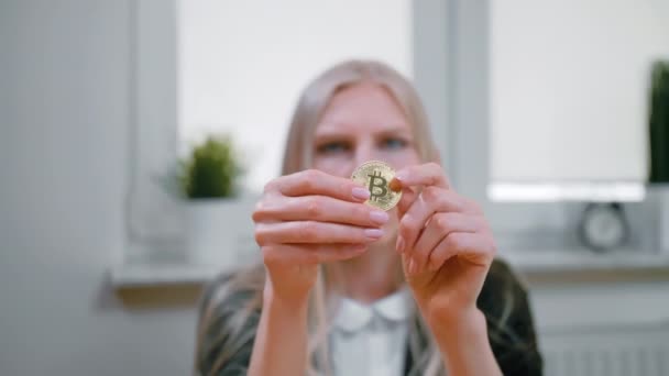 Femme étudiant Bitcoin dans les mains. Vue rapprochée des mains de la jeune femme blonde dans des vêtements élégants assis à la fenêtre et regardant attentivement tenu dans les mains Bitcoin sur fond flou . — Video