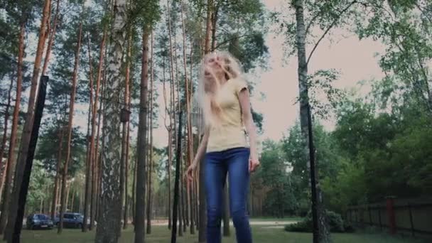 Glad kvinna hoppar på studsmatta. Vacker ung blond barfota kvinna i ljus sommar skjorta och jeans tittar på kameran och ler hoppa på stor studsmatta med höga träd runt. — Stockvideo