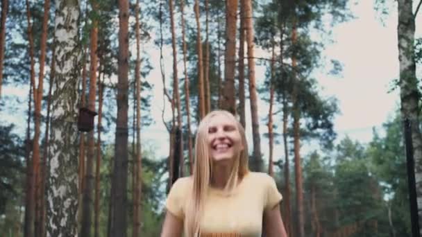 Счастливая женщина прыгает на батуте. Красивая молодая блондинка босиком женщина в светлой летней рубашке и джинсах смотрит в камеру и улыбается прыгая на большом батуте с высокими деревьями вокруг . — стоковое видео