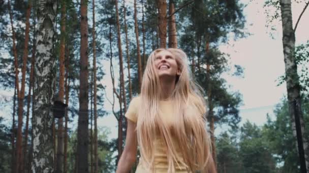 Щаслива жінка стрибає на батуті. Красива молода блондинка босоніжка в легкій літній сорочці і джинсах дивиться на камеру і посміхається стрибати на великому батуті з високими деревами навколо . — стокове відео