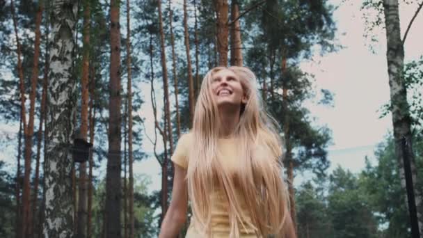 Joyeux saut féminin sur trampoline. Belle jeune femme blonde pieds nus en chemise d'été légère et jeans regardant la caméra et souriant sautant sur un grand trampoline avec de grands arbres autour . — Video